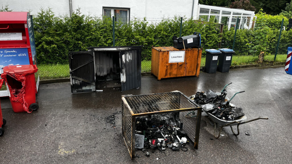Outdoor waste area with multiple bins, a burnt red trash can, and a rusted metal cage filled with burnt debris on a wet pavement.