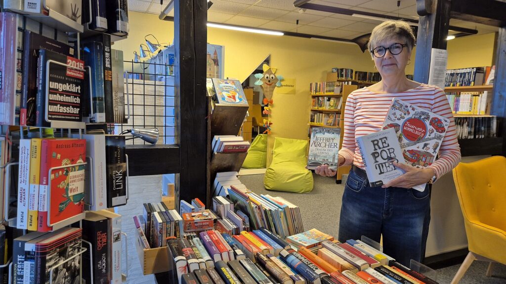 Older woman with short gray hair and glasses in a bookstore, holding several books while standing near shelves of books behind her.