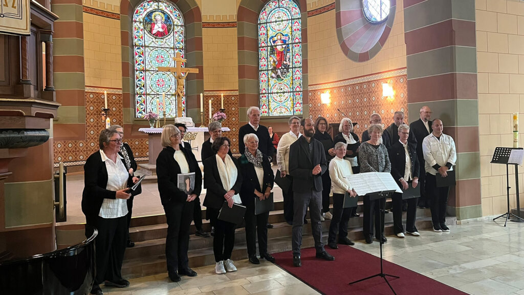A choir of men and women stands in a church pew area, holding folders, facing forward for a performance in front of stained glass windows.