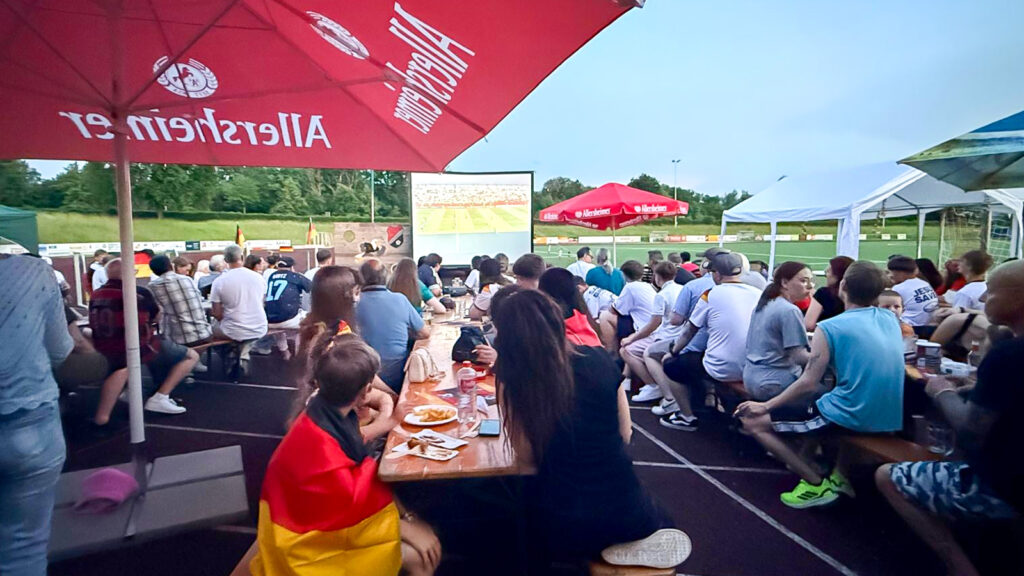 Crowd of people sitting at long picnic tables under red umbrellas, watching a large outdoor football match projected on a screen on a grass field.