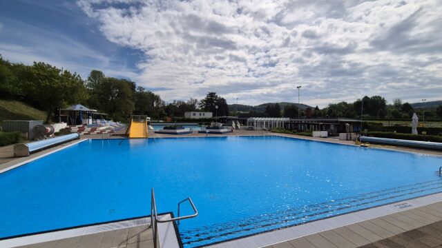 Outdoor pool complex with bright blue water, a yellow slide, and lounge chairs along the deck under a partly cloudy sky.