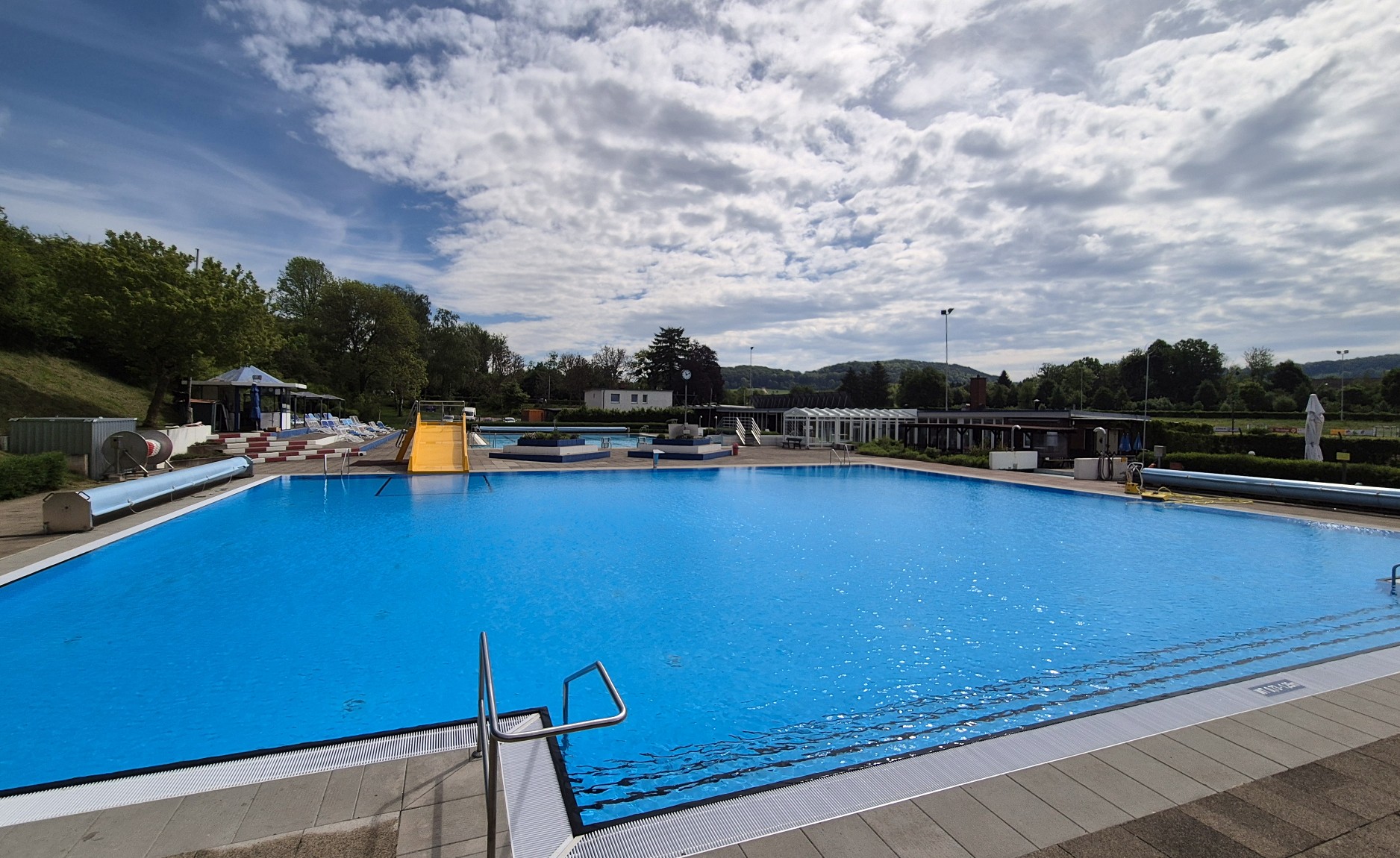Outdoor pool complex with bright blue water, a yellow slide, and lounge chairs along the deck under a partly cloudy sky.
