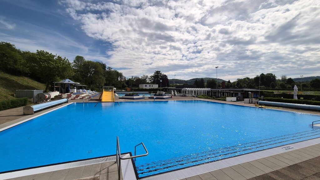 Outdoor public swimming pool with blue water, yellow slide, lounge chairs, and surrounding greenery under a partly cloudy sky.