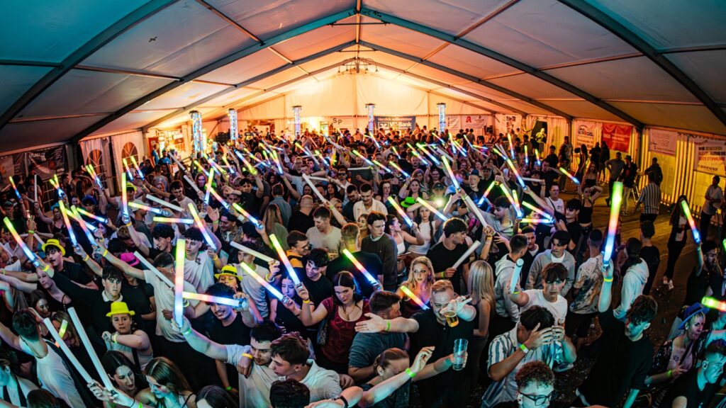 Large crowded indoor party under a tent, people waving colorful glow sticks and dancing.",