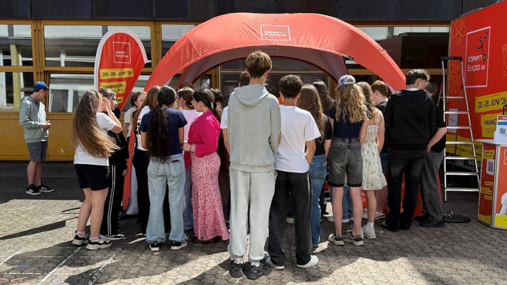 Group of teenagers gathered under a large red canopy at an outdoor expo booth, facing inward and chatting.