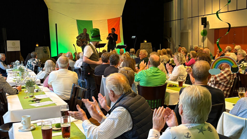 Audience at a banquet hall applauds a live band on stage with Irish flag backdrop and green lighting.