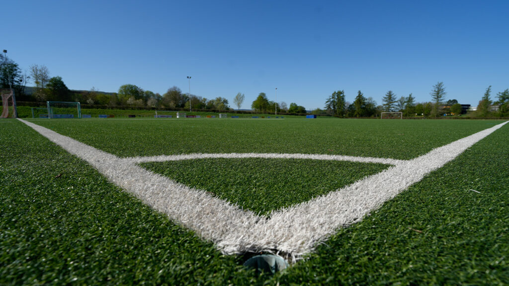 Close-up view of a green artificial turf soccer field corner with white boundary lines converging toward the center, goals visible in the distance under a clear blue sky.