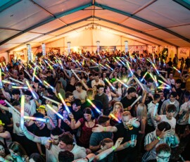 Large crowded indoor party under a tent, people waving colorful glow sticks and dancing.",