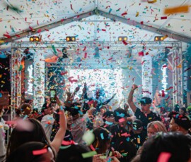 Crowd dancing at a lively indoor party with colorful confetti midair and a stage in the background beneath a tent roof.