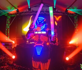 Performer in a glowing LED suit holding illuminated light sticks on a colorful stage with neon beams and confetti on the floor.