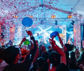 A crowded indoor concert with confetti falling, hands raised, and a brightly lit stage with performers under blue and pink lights.