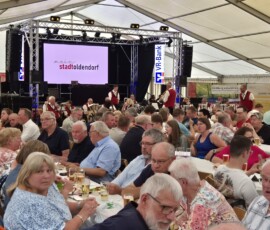Large crowd seated at long tables in a festival tent watching a live band on stage, with banners and a screens displaying 'mein stadtoldendorf' in the background