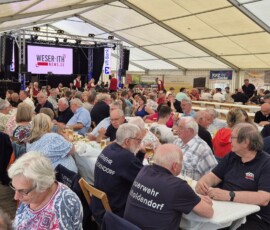 Crowd of mostly older adults seated at long tables inside a large festival tent, facing a stage with a screen and musicians performing.",
