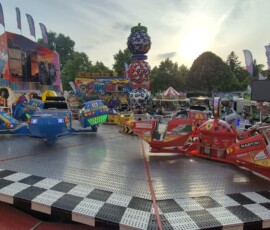 Bumper cars on a circular metal platform at a sunny carnival, with colorful mural walls and stacked decorative spheres.