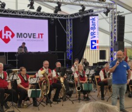Brass band in red vests playing on a stage under a tent, with a man in a blue shirt speaking into a microphone in front of them.