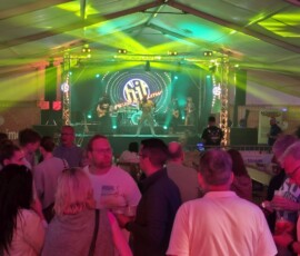 Band performs on a lit stage inside a large tent, with green and yellow spotlights and a crowd watching from tables and standing area.