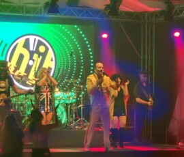 Live band on stage performing under colorful lights; lead singer in a yellow suit with microphone, guitarist on the left, backup singers center, green circular LED backdrop in the background.