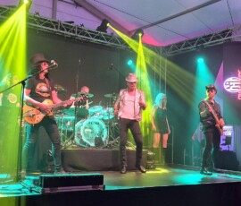 Live rock band performing on a well-lit stage with yellow and green spotlights, guitarist on the left, lead singer center, drummer in the back, and a female backing singer with a male guitarist on the right.