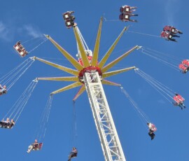 Swing ride with riders in red seats circling a tall central tower against a clear blue sky.