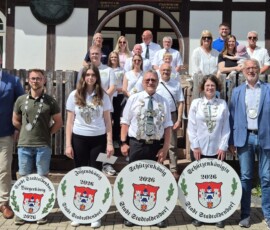 Group of people posing on steps in front of a timber-framed building; several wear ceremonial sashes and large round shields with 'Schützenkönig 2026' and a city crest, indicating a traditional marksmen event.