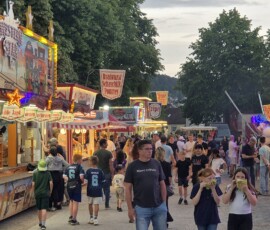 Busy outdoor fair street with colorful food stalls, string lights, and crowds of people shopping and queuing for treats.