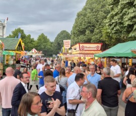 Bustling outdoor carnival scene with colorful game booths, food stalls, and a crowd of people enjoying the fair.